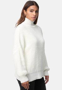 Witte coltrui in een gestructureerde breiwerk, met oversized mouwen en een geribbelde zoom. Draagt zwart broek.