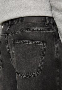 Mustat farkut, joissa on kulunut pinta, viisi taskua ja merkkipaikka nahkavyötäröllä, nähtynä takaa.