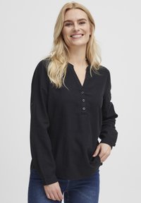 Top nero a maniche lunghe con scollatura a V, patta con cinque bottoni e tessuto morbido e strutturato. Abbinato a jeans in denim blu.