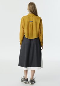 Camisa corta de color mostaza con tela texturizada, combinada con una falda gris oscuro con un dobladillo blanco, llevada con zapatillas deportivas grises.