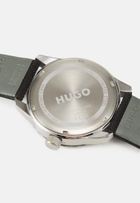 Reloj Hugo con caja de acero inoxidable, correa de cuero gris y grabado detallado en la parte trasera; presenta un botón texturizado para el ajuste.