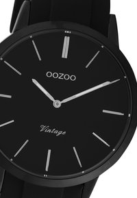 Montre analogique noire avec un cadran noir mat, des index et aiguilles en argent. Présente le nom de la marque "OOZOO" et "Vintage" sur le cadran.