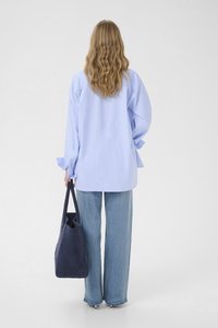 Mujer de cabello rubio ondulado, vestida con una camisa azul claro de gran tamaño, vaqueros de pierna ancha y sosteniendo un bolso tote azul marino, vista de espaldas.