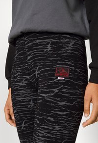 Svarta leggings med en texturerad grå mönster, med en röd logopatch på sidan. En del av en avslappnad outfit med en grå sweatshirt.