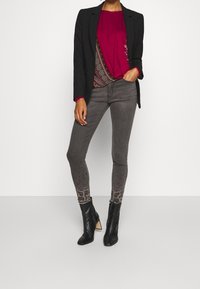 Schwarze Blazer über einem burgunderfarbenen Oberteil, graue gemusterte Skinny-Jeans mit strukturierten Akzenten und schwarze Absatzstiefeletten.