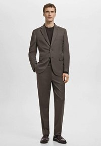 Traje marrón a medida con un blazer de un solo pecho y pantalones a juego. La tela parece suave. Camisa negra debajo. Zapatos oscuros completan el conjunto.