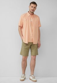 Korte mouwen, licht perzik kleurige button-up shirt over een bijpassend T-shirt, gecombineerd met olijfgroene shorts en witte sneakers met beige accenten.