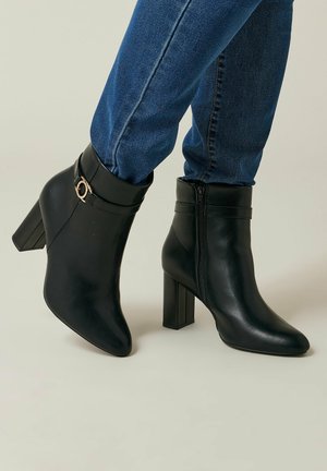 Schwarze Knöchelstiefel aus glattem Leder mit einer spitzen Zehe, einem Reißverschluss und einem Blockabsatz. Ein goldener Akzent ziert den Knöchelriemen.