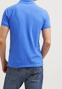 Uomo che indossa una polo a maniche corte blu brillante e jeans blu, mostrato di spalle contro uno sfondo chiaro e uniforme.