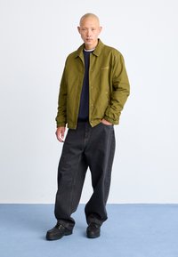 Veste légère vert olive avec un design à col, doublure intérieure bleu marine, associée à un jean noir ample et des chaussures noires.