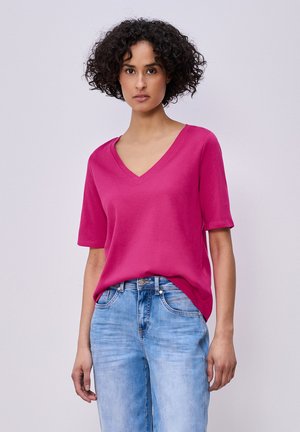 Femme aux cheveux bouclés portant un t-shirt rose vif à manches courtes et col en V, ainsi qu'un jean en denim bleu clair, debout devant un fond uni.
