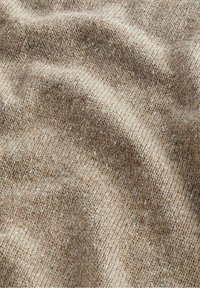 Beige gestrickter Stoff mit strukturierter Oberfläche, der eine Mischung aus hellen und dunklen Fasern zeigt und ein dezentes meliertes Muster erzeugt.