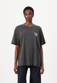 T-shirt oversize grigio scuro, maniche corte, scollo rotondo, con un logo a forma di cuore colorato sul lato sinistro del petto. Materiale in cotone morbido.