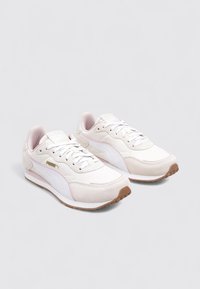 Un par de zapatillas Puma blancas y rosa claro con cordones blancos y suelas de goma marrón, vistas en ángulo sobre un fondo liso.
