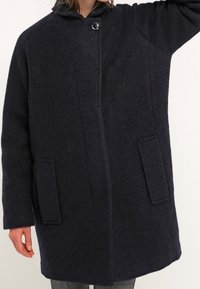 Manteau en laine bleu marine avec col montant, fermeture à boutons sur le devant et deux grandes poches plaquées. Présente une coupe ample et un tissu texturé.