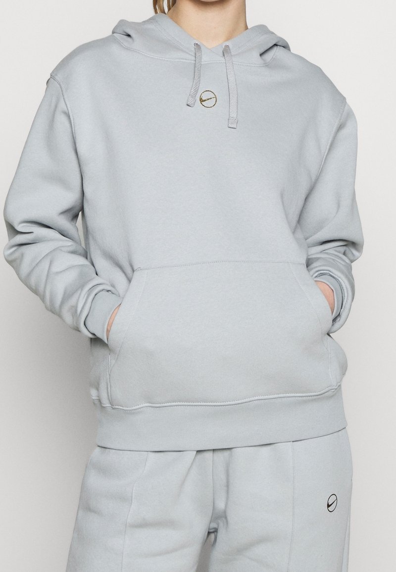 Ljusgrå hoodie tillverkad av mjukt tyg med en framficka, dragsko i huvan och en liten gulddetalj med logotyp på bröstet.