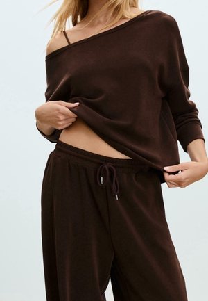 Frau trägt dunkelbraune, weite Hose und schulterfreies Langarm-Oberteil, hebt das Oberteil leicht an, um Taille und schwarzen BH-Träger zu zeigen.