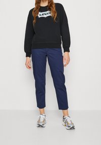 Personne portant un sweat-shirt noir Levi's, un pantalon cropped bleu marine et des baskets blanches avec des accents orange et argent, se tenant sur un fond neutre.