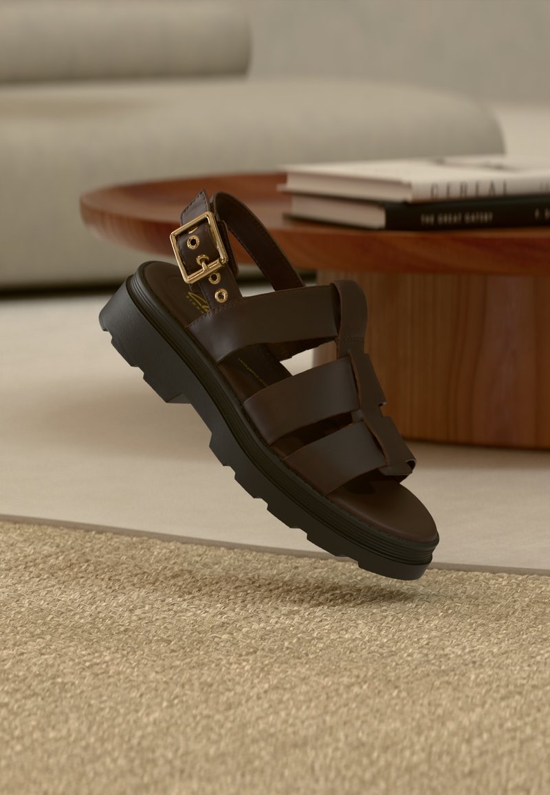 Σκούρο καφέ δερμάτινο σανδάλι με μαύρη χοντρή σόλα και χρυσή αγκράφα, τοποθετημένο πάνω από μπεζ χαλί κοντά σε ξύλινο τραπέζι με στοιβαγμένα βιβλία.