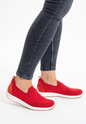 Legs in fitted dark gray jeans wearing bright red slip-on athletic shoes with white soles and brown tread on a white background.