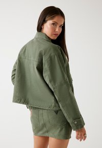 Jachetă scurtă verde măsliniu, confecționată dintr-un material durabil, cu guler, design pătrățos și manșete cu nasturi, asociată cu o fustă asortată.