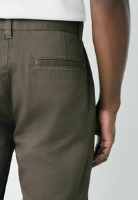 Pantaloni verde închis din materiale netede, având un buzunar posterior și o croială ajustată, prezentați dintr-un unghi lateral, cu mâna aşezată pe șold.