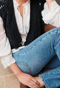 Gilet nero con frange indossato sopra una camicetta bianca con balze, abbinato a jeans blu chiaro con dettagli in metallo lungo il lato.