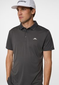 Mörkgrå polo skjorta med en slät textur, treknappsknäppning och en vit logotyp på vänster bröst. Klassisk kragsdesign.