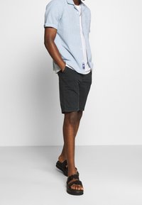 Košile s krátkým rukávem, světle modrý pruhovaný vzor, černé kraťasy a černé sandály. Košile má límec a kraťasy mají postranní kapsy.