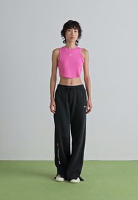 Dünne Person steht, trägt ein pinkes ärmelloses Crop-Top, lockere schwarze Sweatpants mit Logos und weiße Sneaker auf einem grünen Boden.
