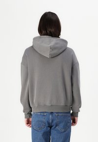 Sweatshirt griș cu glugă, cu o croială lejeră, manșete și tiv din bumbac ribetat. Purtați cu blugi albaștri, prezentând o textură fină și un design casual.