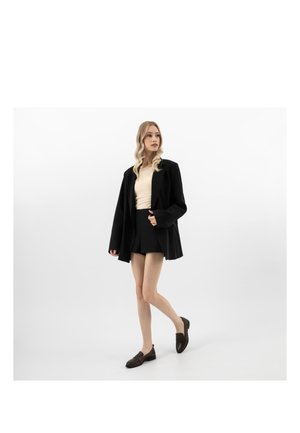 Femme aux cheveux longs et blonds portant un haut beige, un blazer noir surdimensionné, un short noir et des mocassins foncés, debout devant un fond blanc uni.