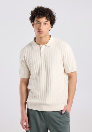 Junger Mann mit lockigem Haar, der ein cremefarbenes, kurzärmliges, gestricktes Polohemd und dunkelgrüne Hosen trägt, steht mit einer Hand in der Hosentasche.