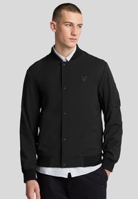 Giacca bomber nera con colletto e polsini a coste, con un logo discreto. Texture liscia e chiusura con bottoni, indossata sopra una camicia con colletto.