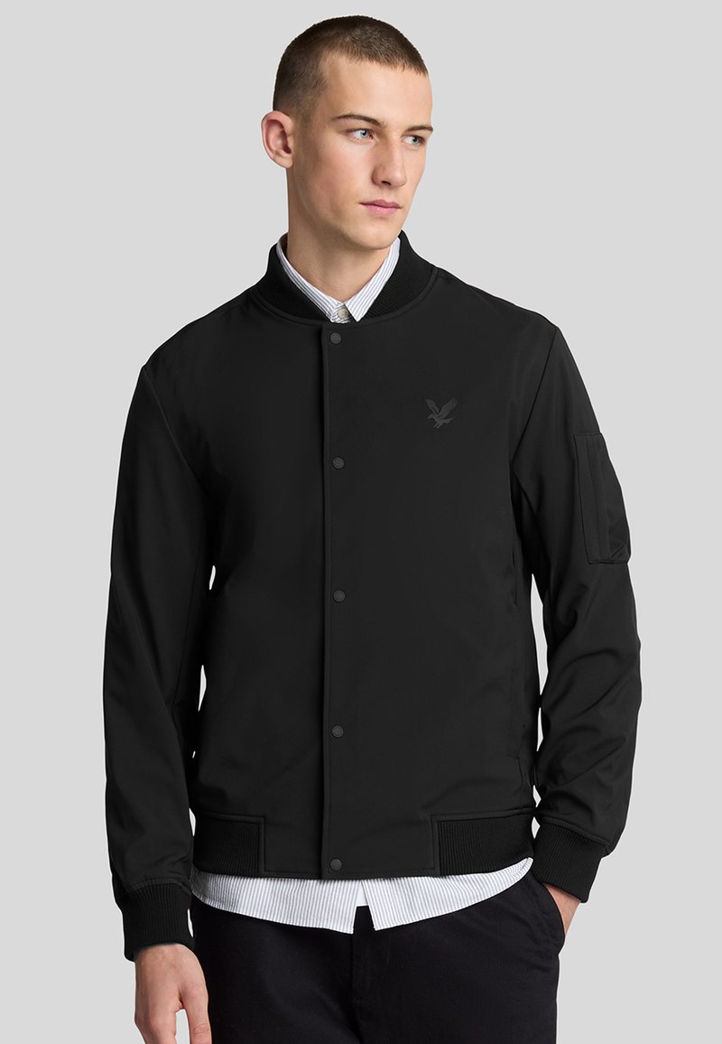 Giacca bomber nera con colletto e polsini a coste, con un logo discreto. Texture liscia e chiusura con bottoni, indossata sopra una camicia con colletto.