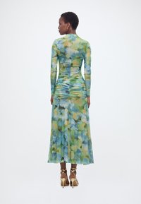 Γυναίκα με την πλάτη γυρισμένη, φοράει ένα μακρυμάνικο φόρεμα με πράσινα και μπλε άνθη, με ραβδώσεις στη μέση και χρυσά ψηλοτάκουνα σανδάλια, σε λευκό φόντο.