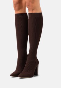 Bottes brunes à côtes, montantes jusqu'au genou, avec un bout pointu et un talon empilé, fabriquées dans un tissu texturé qui épouser la jambe.