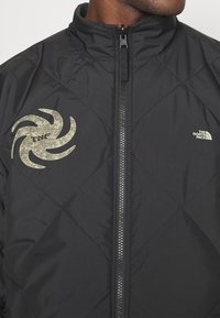 Veste matelassée noire avec fermeture éclair frontale, avec un logo TNF circulaire clair sur la poitrine gauche et un logo The North Face sur la poitrine droite.
