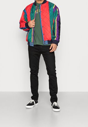 Kleurrijke doorgestikte bomberjack met rode, groene, blauwe en paarse strepen; geribbelde boorden en kraag; gedragen over een groen shirt met zwarte spijkerbroek.