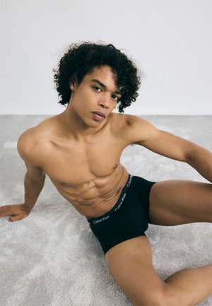 Mustat boxerit, joissa on Calvin Kleinin logo vyötäröllä, yhdistettynä päivettyneeseen ihoon ja määriteltyyn lihasrakenteeseen, istuu vaaleanharmaalla pinnalla.