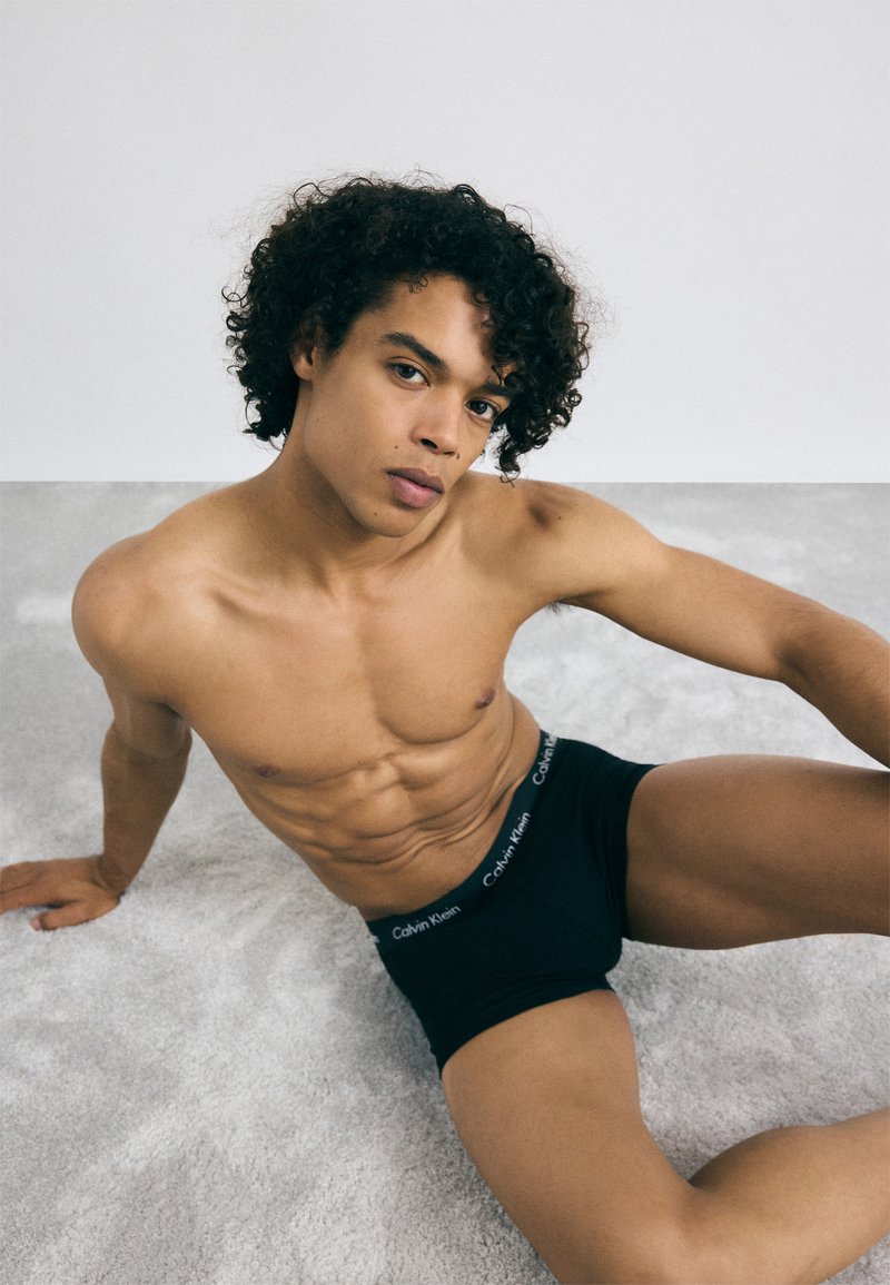 Boxeri negri, cu un logo Calvin Klein pe talie, asortați cu o piele bronzată și o structură musculară bine definită, așezați pe o suprafață gri deschis.