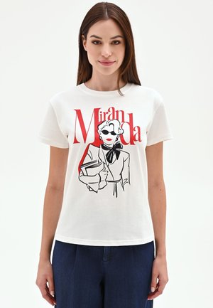 Donna che indossa una maglietta bianca con una grafica nero-rossa di una donna elegante con occhiali da sole che tiene dei libri e la scritta "Miranda" sul davanti.