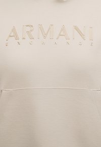 Tissu beige avec texte beige brodé en relief "ARMANI EXCHANGE" sur le devant, au-dessus d'une couture horizontale et d'une ouverture de poche.