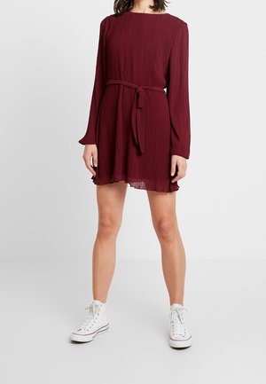 Femme portant une robe plissée bordeaux à manches longues avec une ceinture à la taille et des baskets montantes blanches, debout devant un fond blanc uni.