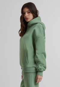 Hoodie vert clair en tissu doux, avec une poche avant, des poignets côtelés et une capuche à cordon de serrage. Logo blanc sur la poitrine.