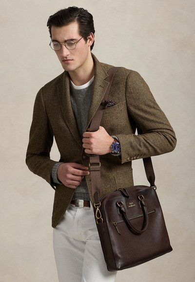 Man med glasögon, brun kavaj, grå tröja, vita byxor, blå klocka, bär en mörkbrun läderportfölj med axelrem.