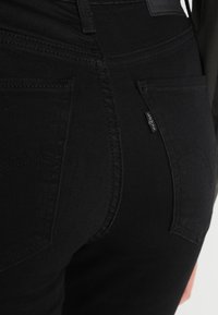 Jeans de mezclilla negra con un diseño ajustado, que cuentan con dos bolsillos traseros y detalles de costura sutiles. Etiqueta visible en el lateral.
