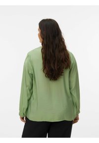 Chemise vert clair à manches longues en matériau doux, avec une coupe décontractée et des poignets boutonnés, vue de dos. Un pantalon noir est visible.