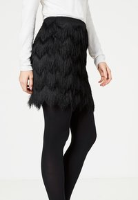 Svart kjol med en texturerad fransdesign, har en scalloped nederkant och ett brett elastiskt midjeband, ihop med svarta strumpbyxor.
