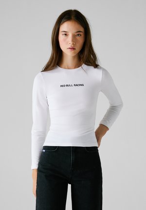 Haut à manches longues blanc avec une coupe ajustée, arborant le texte "RED BULL RACING" en noir sur la poitrine. Porté avec un jean sombre.